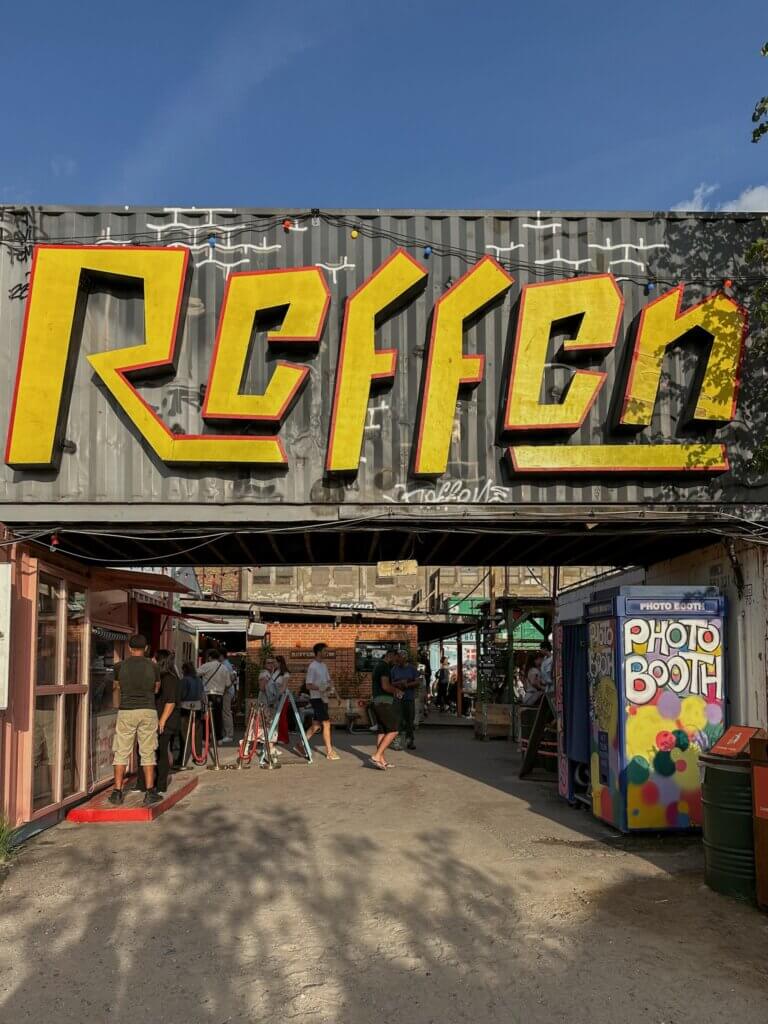 Large yellow letters spelling "REFFEN" mounted on a grey shipping container entrance above a bustling outdoor street food market, with a colorful photo booth on the right in Copenhagen.