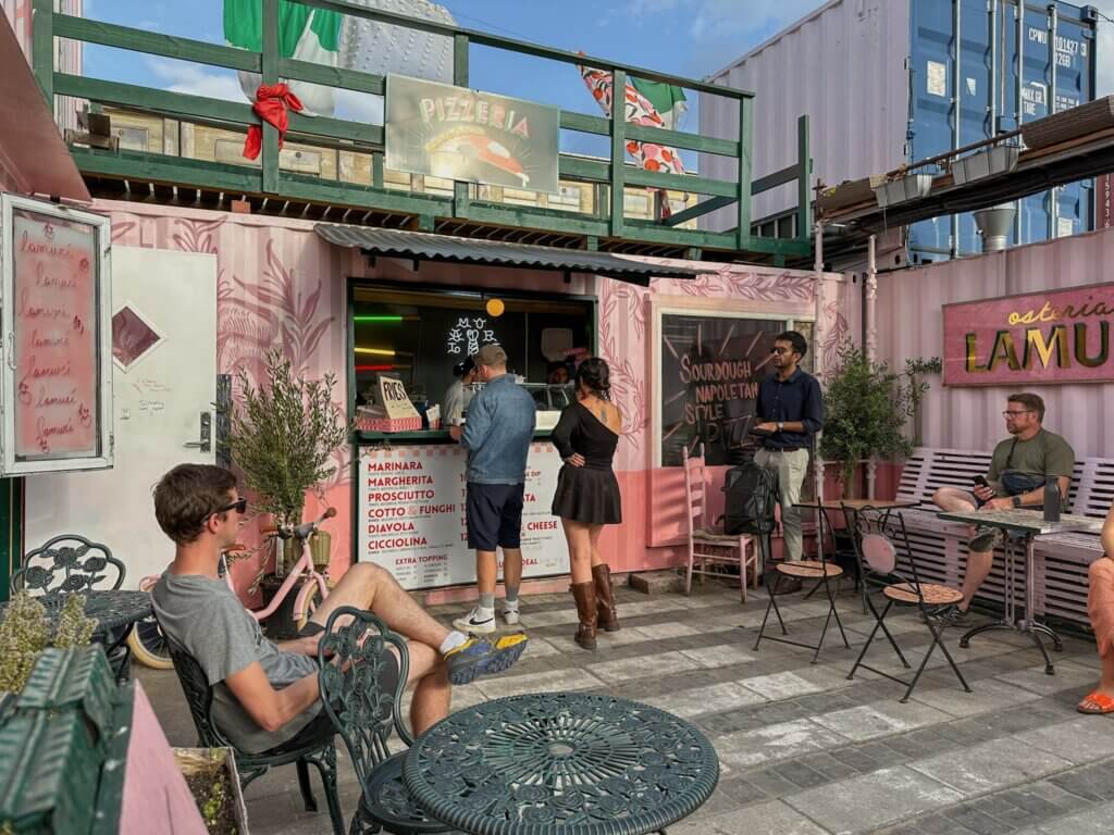 People hanging out at an outdoor pink shipping container pizzeria stall called Osteria Lamuci, with small metal patio tables and seating at Reffen Street Food Market in Copenhagen.
