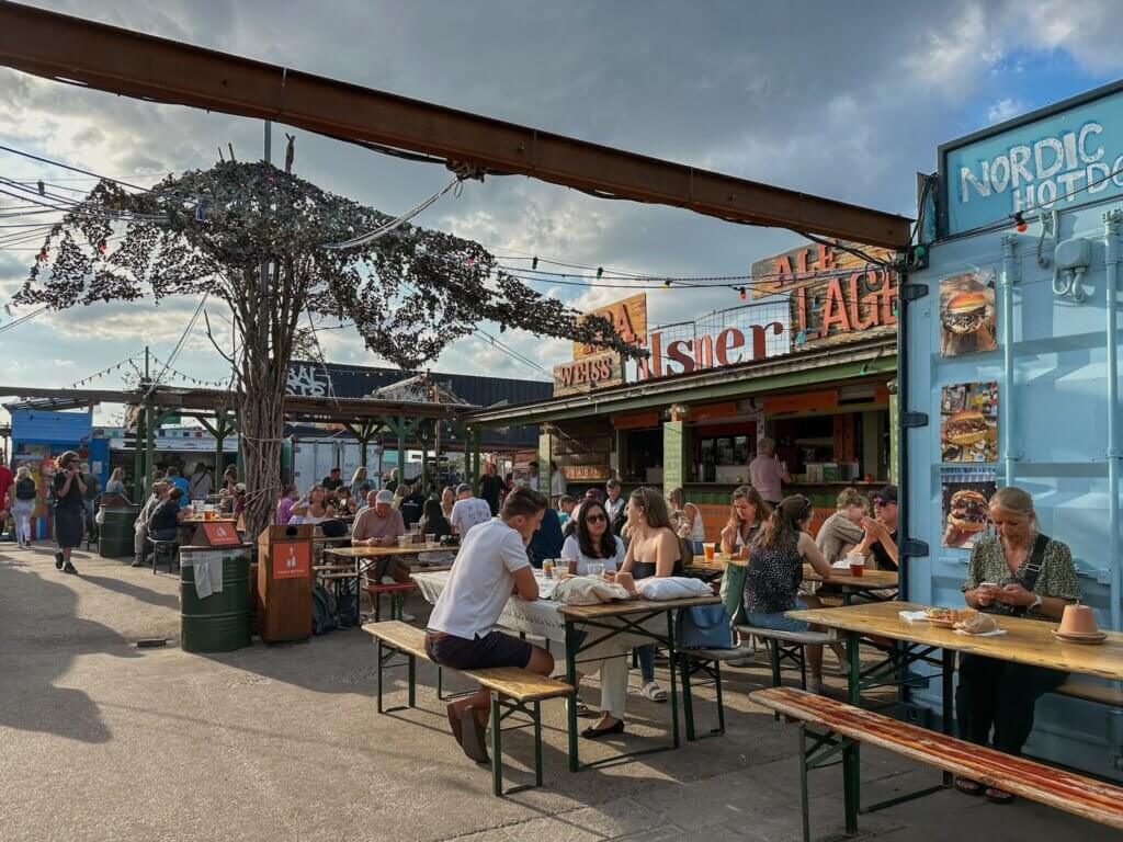 People enjoying food and drinks at outdoor wooden picnic tables at Reffen Street Food Market, surrounded by shipping container stalls, string lights, and signs reading "Nordic Hotdog" and "Pilsner" in Copenhagen.