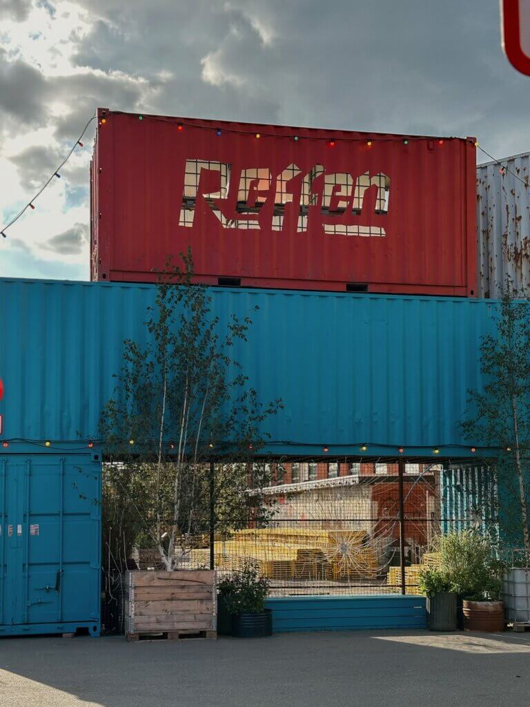 A red shipping container featuring the cut-out word "Reffen" stacked on top of a blue shipping container frame with string lights against a cloudy sky in Copenhagen.