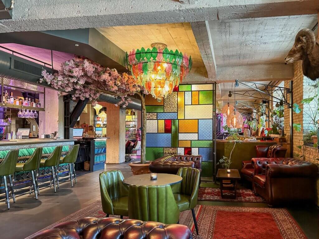 Eclectic hotel lounge interior featuring green leather chairs, dark brown tufted chesterfield sofas, a vibrant glass leaf chandelier, stained-glass divider, and a pink cherry blossom tree in Copenhagen.