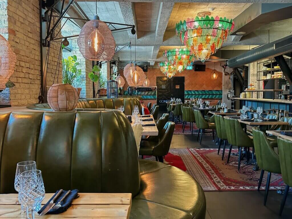 Stylish hotel restaurant interior featuring green leather booth seating, exposed brick walls, woven pendant lights, and colorful glass leaf chandeliers over laid-out dining tables.