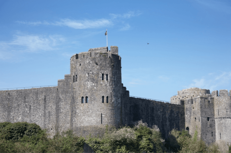 A large , historic stone castle, Pembroke Castle, featuring a massive round keep flying a flag under a blue sky. It is a must visit on a dog-friendly break in Wales.
