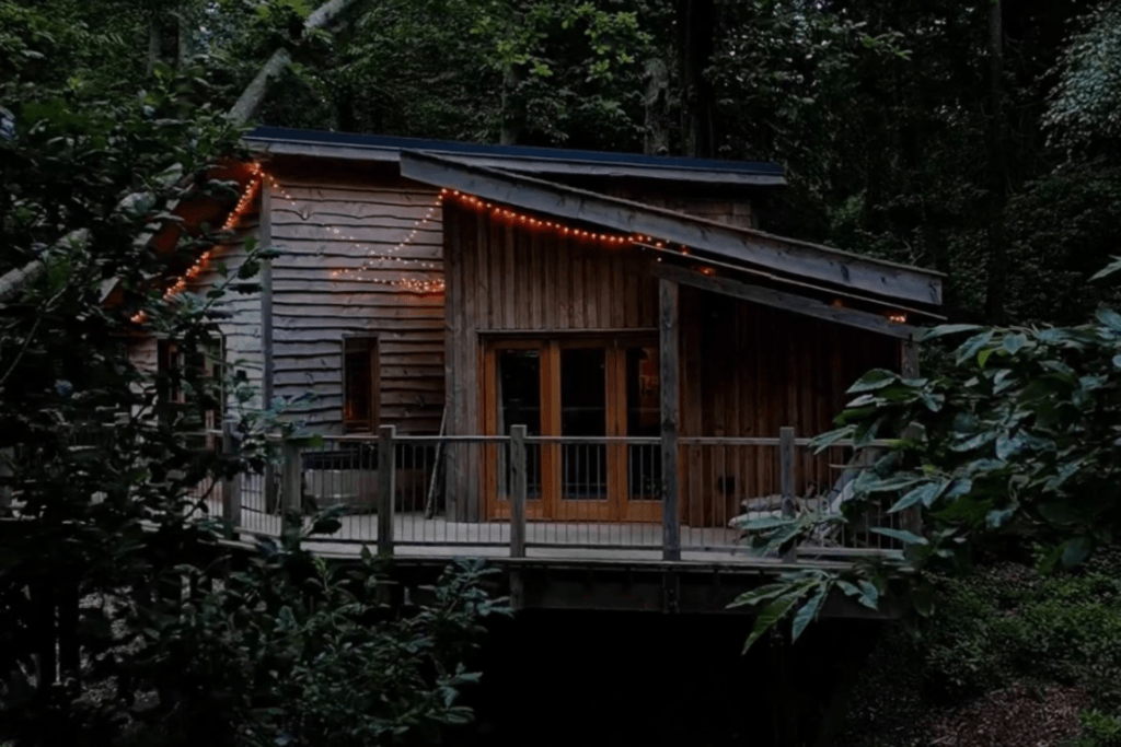An evening view of a rustic wooden treehouse nestled among dense green trees, featuring a wooden deck with a railing and warm string lights glowing along the edge of its slanted roofline.