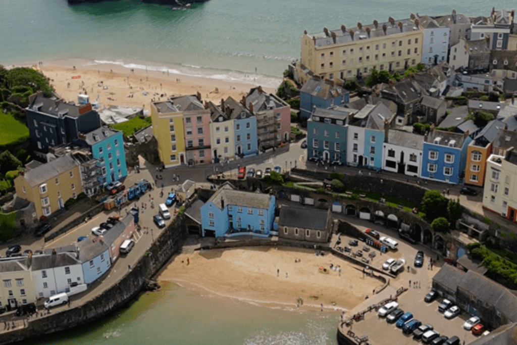 An aerial, high-angle view of Tenby, a coastal town in Wales featuring rows of brightly coloured pastel buildings overlooking a sandy beach cove. In the foreground, a curved stone sea wall encloses a small harbour beach with a central blue building and a stone structure. Above the harbour, streets lined with yellow, pink, blue, and white multi-story houses sweep around the landscape. In the background, a second, larger sandy beach extends along the calm, greenish-blue sea under bright daylight.
